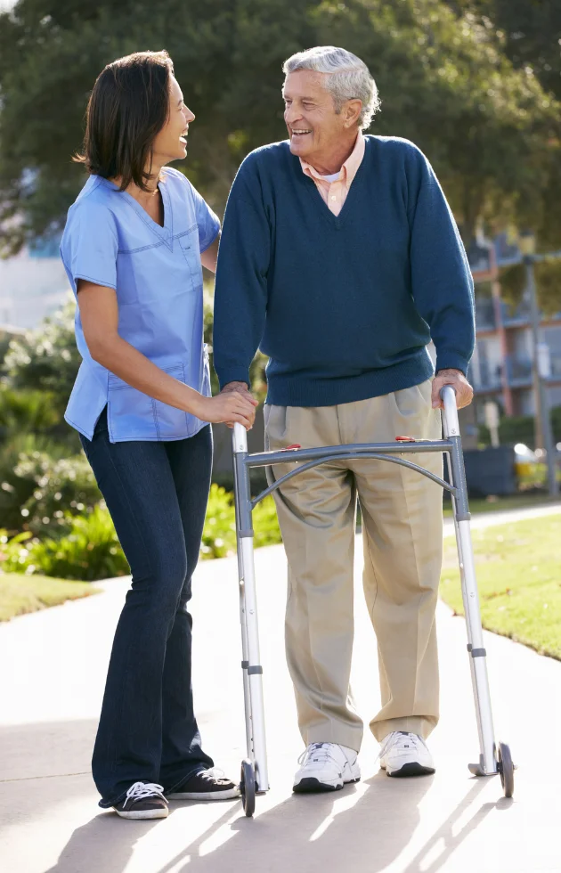 Female carer walking alongside an older adult using a walking frame outdoors.