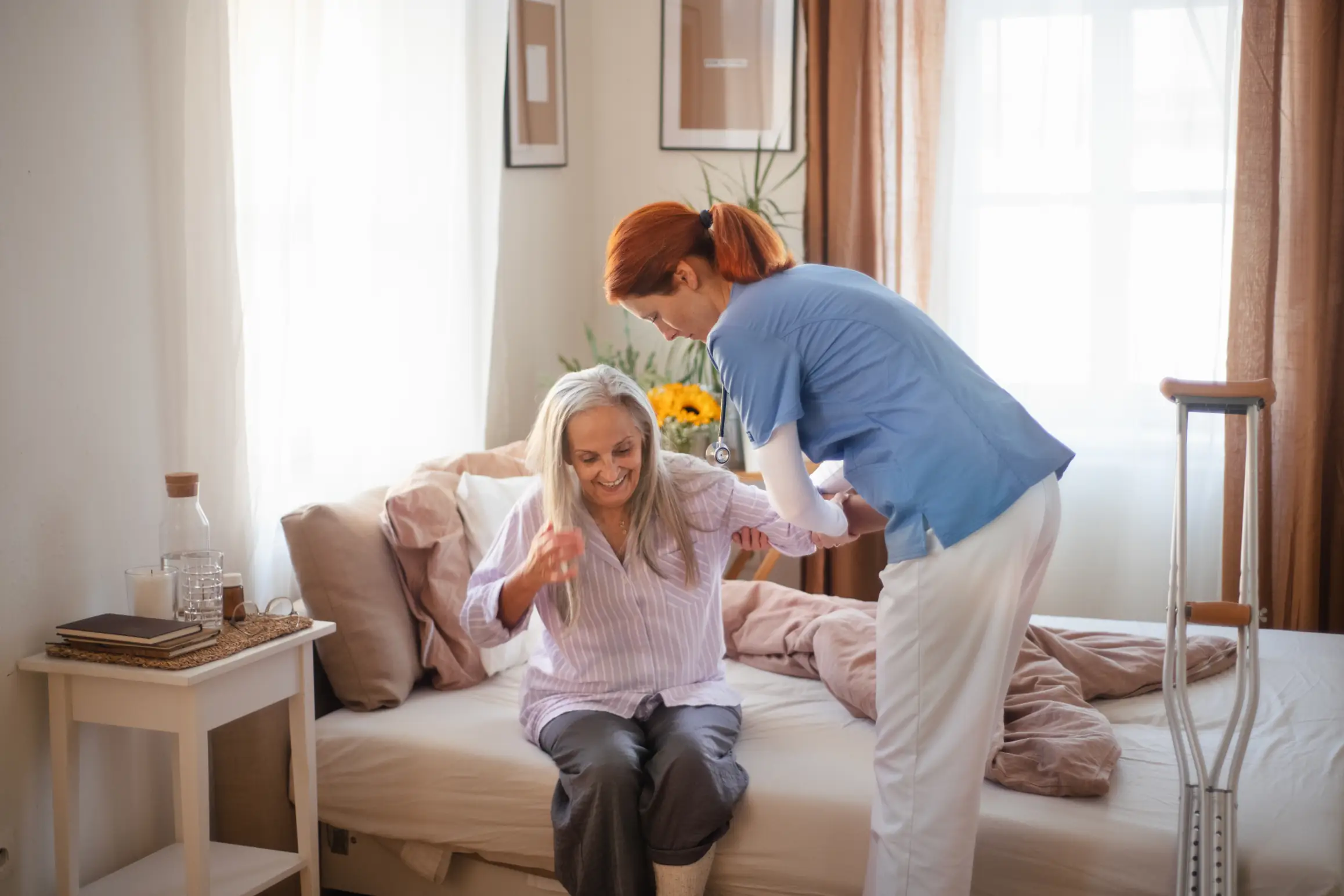 Experienced live-in nurse in Truro and Swindon aiding a smiling senior woman to get out of bed, offering post-injury recovery support.