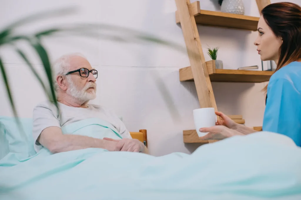 Female nurse offering a drink to an elderly male patient in bed, part of palliative care in Truro or Swindon.