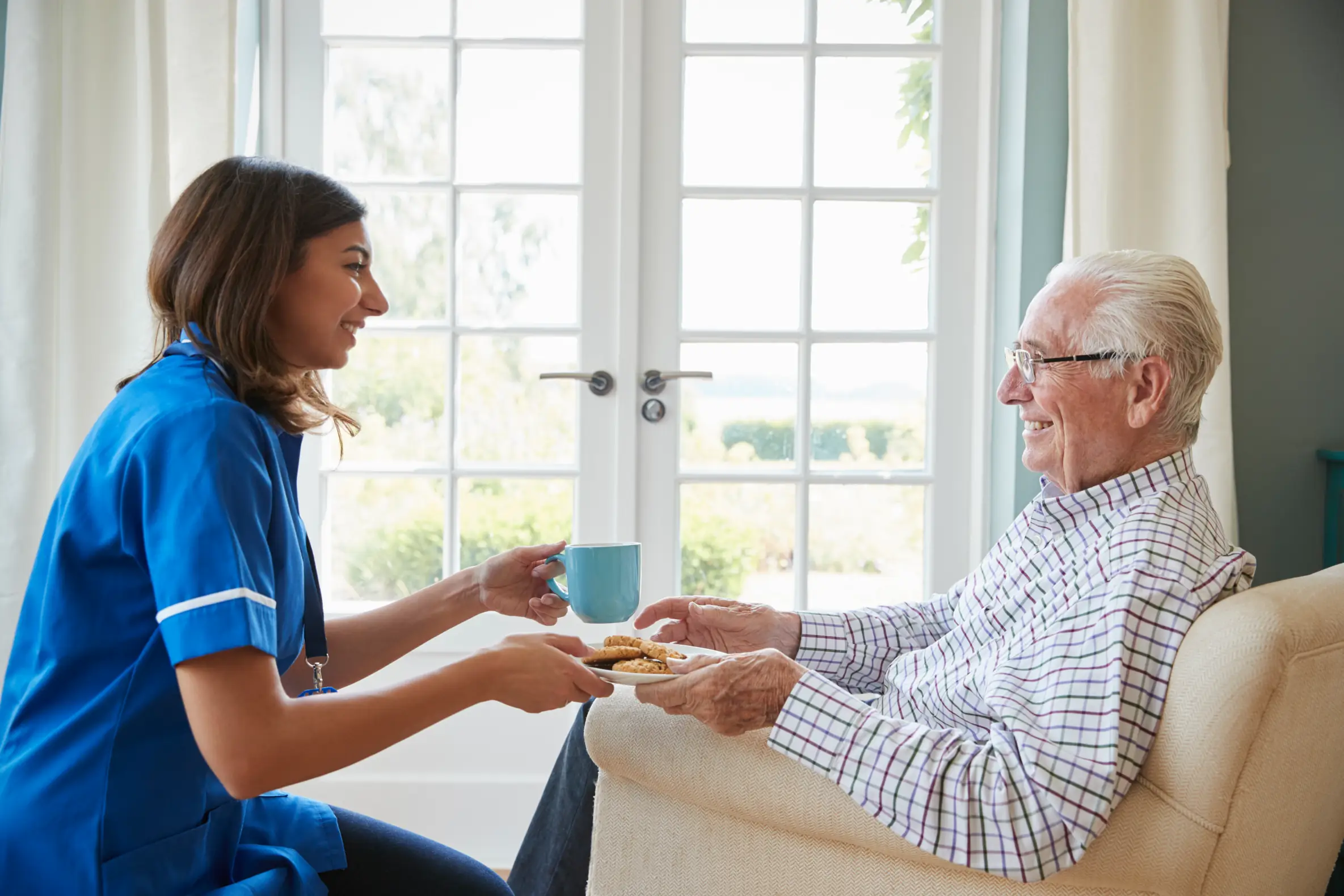 Friendly live-in carer in Truro and Swindon serving tea and biscuits to a cheerful senior man in his armchair, showcasing companionship.