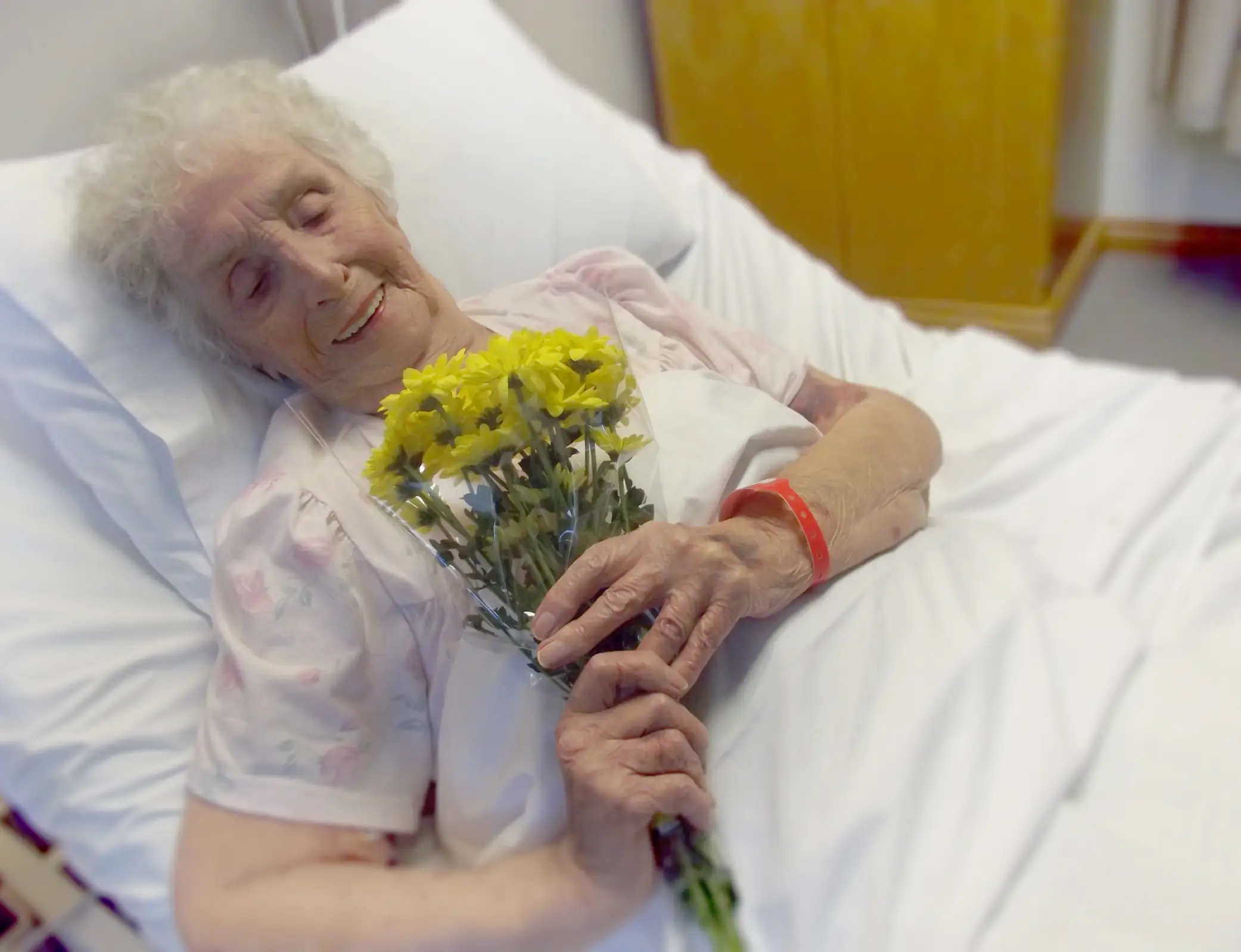 Elderly woman in bed smiling, holding yellow flowers, receiving palliative care in Truro or Swindon.