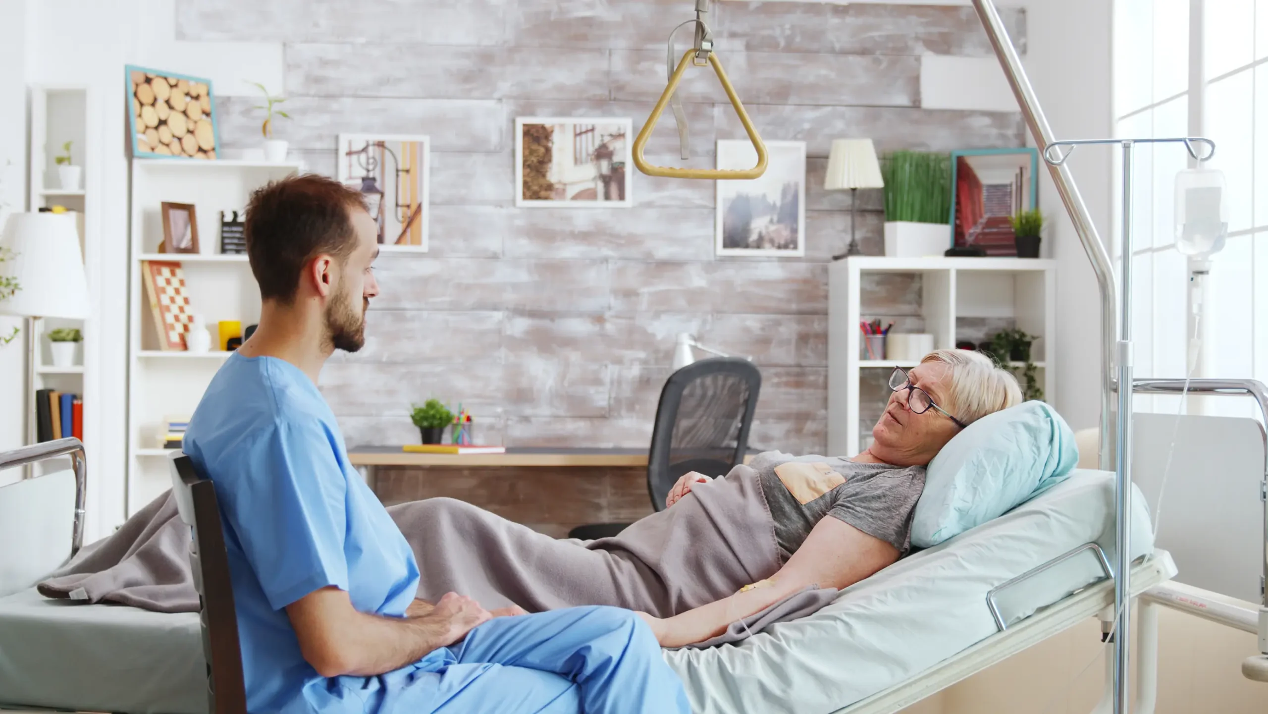 Male carer attentively listening to an elderly woman in a hospital bed, palliative care services in Truro and Swindon.